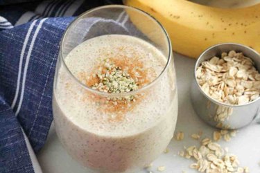 banana chai smoothie in a glass surrounded by oats and banana on a white surface