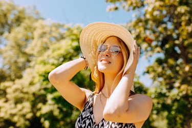 Close view of a person with long blonde hair standing in the sun wearing a big sun hat and sunglasses, to represent limiting sun exposure with lupus