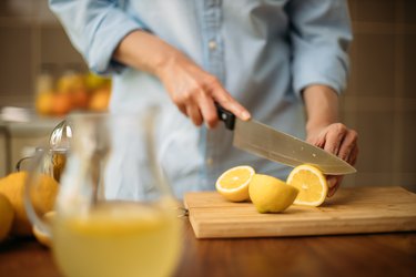 Close up of person in a blue shirt cutting lemons on a wooden cutting board next to a pitcher of lemon juice in the kitchen.