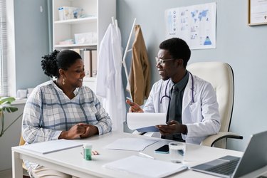 a smiling person wearing a button-down flannel shirt speaks to a young doctor wearing a white lab coat and stethoscope in a sunny office about the health benefits of breast reduction surgery