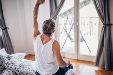 a rear view of an older adult wearing a white tank top and blue flannel pajama pants stretching while sitting on the side of a bed in the morning