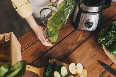 overhead view of a person pouring a green smoothie from a blender into a glass, with fruits and veggies nearby on a cutting board