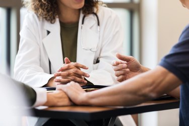 Couple holds hands while husband gestures and speaks to doctor