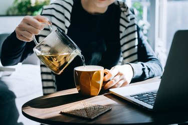 Close up of hands pouring a cup of medicine ball tea from a stylish transparent teapot into a cup.