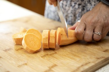 Close-up of female hands cutting a peeled renal diet sweet potato into slices with a kitchen knife on a wooden chopping board