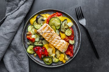 an overhead photo of a roasted salmon filet on top of vegetables like zucchini and yellow summer squash on a black plate next to a black fork and gray cloth napkin