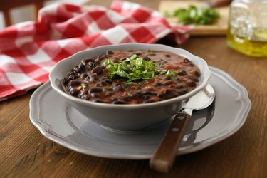 Black bean soup in a gray bowl with fresh chopped parsley on top on a gray plate on wooden table.