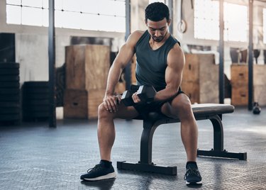 Person wearing a black tank top and shorts performing a biceps curl on a bench at the gym.