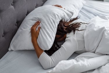 Stressed woman covering head under pillow lying in bed with hangover