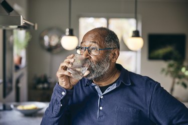 Older adult wearing glasses and a blue long sleeve shirt drinking water at their kitchen sink