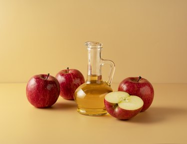 Apple vinegar in a glass container surrounded by apples on a beige background