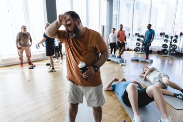 Exhausted older person with a beard and short hair sweating through their orange t-shirt taking break with friends after group training in gym
