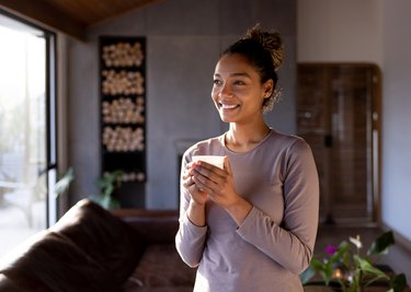 a smiling person with black curly hair in a bun holding a cup of hot chocolate in their living room