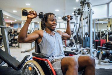 Person in wheelchair working upper-body muscles with dumbbells in a gym.