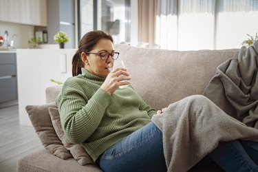 a person wearing a green turtleneck sweater and jeans sits on the couch with a blanket drinking a glass of water