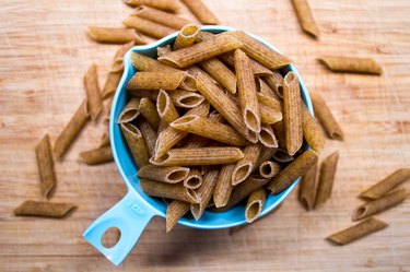 Brown whole-wheat pasta spilling into a measuring cup on wooden table