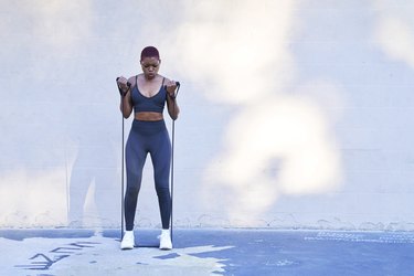 person with short dyed hair wearing a matching sports bra and leggings set doing a resistance band arm workout outdoors on a sunny day