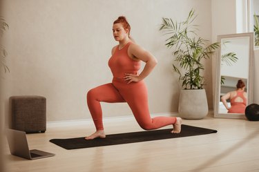 woman dressed in coral workout outfit doing a half-kneeling hip flexor stretch on a black yoga mat at home