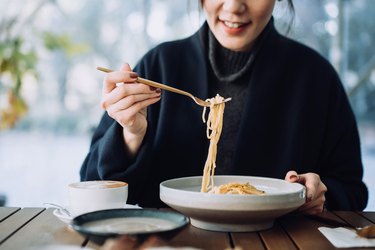 woman sitting at wooden table eating noodles with gluten from white bowl