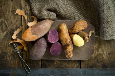 Different types of sweet potatoes partially peeled on wooden cutting board over wooden background.