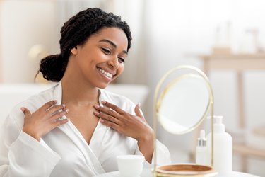 Beautiful Black woman looking at face and neck in mirror and touching décolleté area