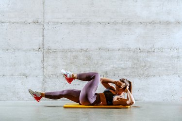 Young strong muscular caucasian sportswoman in sportswear with ponytail lying on the mat and doing cycling cross crunches.