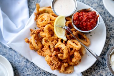 A Plate Of Deep Fried Squid or Calamari Cajun Style, Served With A Side Of Freshly Made Garlic Aioli Sauce And Cocktail Sauce, Fresh Lemon Wedges On The Side