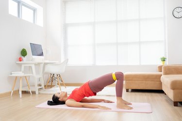 woman doing a resistance band glute bridge on a pink yoga mat in her living room