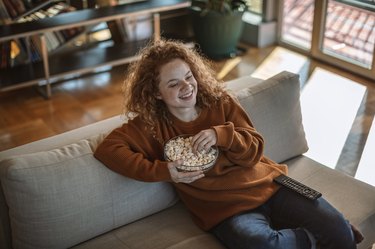 Person with red curly hair and an orange sweater sitting on the couch, eating popcorn from a bowl, as a diabetes-friendly snack