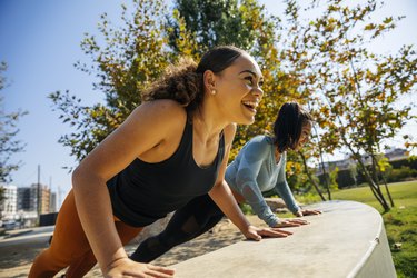 Cheerful people doing push-ups, an exercise for strong, sculpted arms, on retaining wall at park