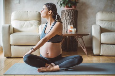 Pregnant woman in a dusty blue sports bra and leggings doing a seated spinal twist yoga pose on a blue yoga mat in the living room, two white leather recliners in the background