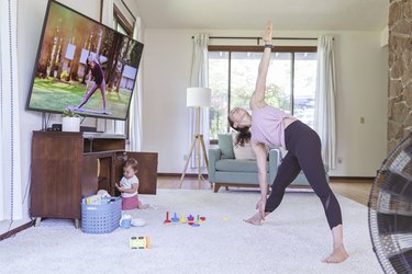 woman doing triangle pose in her living room in front of the tv to a beginner workout dvd, baby playing in background