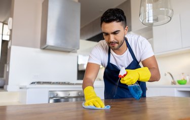 Close up of a man wearing yellow rubber gloves, spraying disinfectant on a kitchen counter and wiping it.