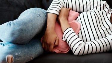 a person in a striped shirt and jeans using a heating pad for cramps while lying on a gray couch