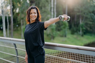 Active energized sportswoman stretches arms with dumbbell, poses on bridge over nature background with sunrise in morning, dressed in active wear, looks into distance, works on training biceps