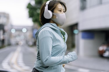 Asian woman running outdoors in a turquoise hoodie with a gray face mask and a white pair of headphones