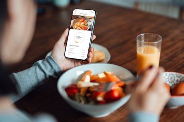Close-up of a person using mobile app to track nutrition and count calories with smartphone while eating fresh fruit breakfast