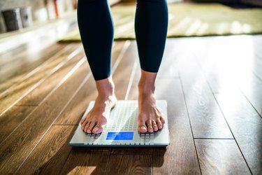 A barefoot person wearing black leggings weighing themselves on a glass scale in their living room.