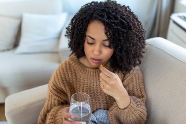 a person with natural hair wearing a light brown chunky sweater sitting on a beige couch and holding a biotin supplement and a glass of water