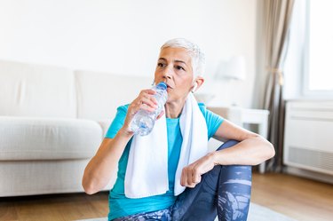 Older adult woman in workout clothes sitting on the floor in front of the sofa drinking water after a workout