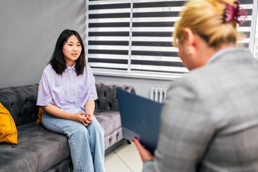 一个十几岁的亚洲女孩坐在灰色的沙发上和治疗师谈论使用心理评估的局限性