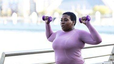 African-American woman in purple, long-sleeve workout top lifting purple hand weights (1 to 2 pounds), doing sciatica exercises outdoors in front of a blurred-out city background