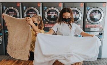 two friends wearing face masks fold laundry together in a laundromat in front of gray washing machines and dryers