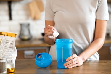 a close up of the torso of a person wearing a white t-shirt scooping whey protein powder into a blue protein shake bottle