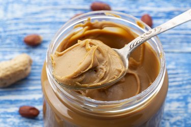 Peanut butter in an open jar and peanuts in the skin are scattered on the blue table.