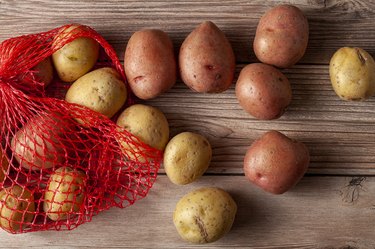 Flat lay  close up image featuring a red mesh potato sack with pink and yellow raw organic potatoes on wooden background