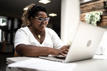 a person with a ponytail wearing a white shirt and black glasses using a laptop working from home