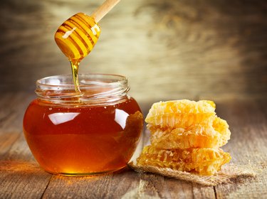 jar of honey, a food high in glucose, with a honey dipper drizzling honey, with honeycomb on a wooden table