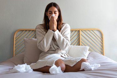 a person with long brown hair wearing a white sweater and sitting in bed while blowing their nose and nursing a cold headache