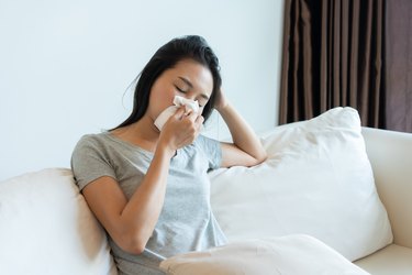 Close up of a person with shoulder length brown hair wearing a gray t-shirt sitting on a cream colored couch blowing her nose on a tissue.